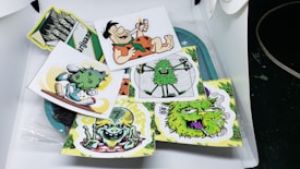 A collection of cartoonish and vibrant stickers featuring various humorous and playful characters, including a caveman with a cigar, smiling green creatures, and quirky designs. Each sticker is colorful and arranged in a casual, overlapping manner on a surface.