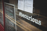 A glass window displays the hashtag '#becurious' in bold white letters. The reflection on the window shows a street with road markings. A chalkboard with handwritten notes and a printed paper with text are visible on the interior of the establishment, indicating a community or event space.