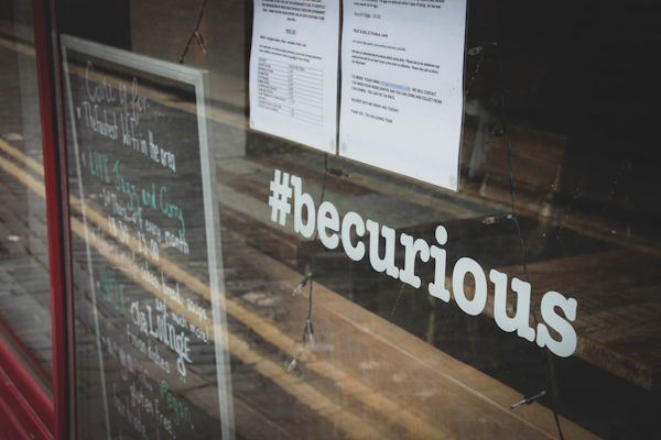 A glass window displays the hashtag '#becurious' in bold white letters. The reflection on the window shows a street with road markings. A chalkboard with handwritten notes and a printed paper with text are visible on the interior of the establishment, indicating a community or event space.