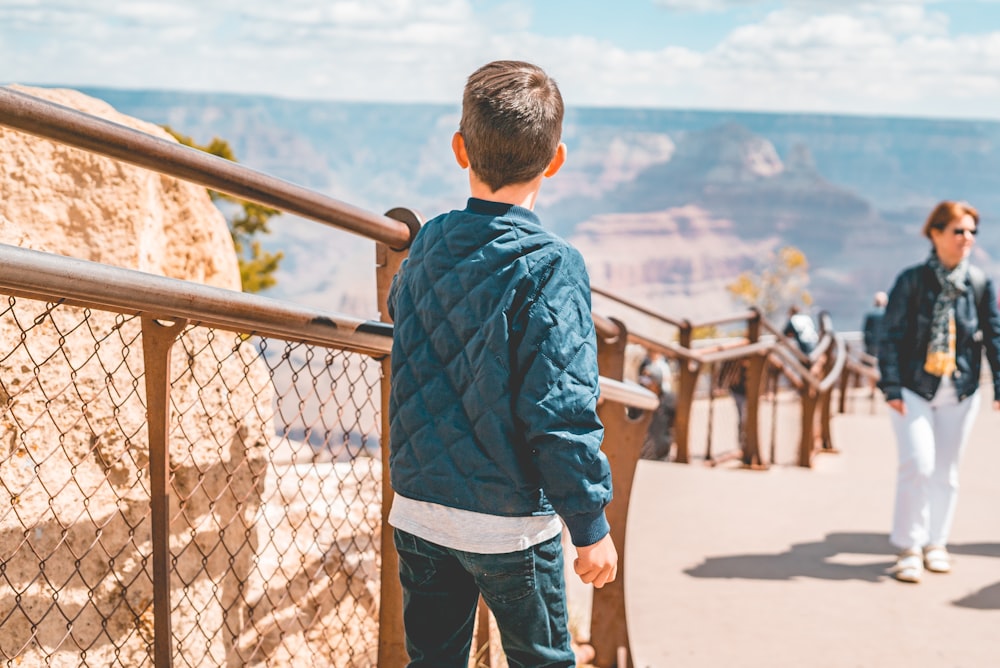 boy in blue and red jacket standing on brown wooden bridge during daytime