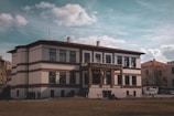 A large, historic building with a two-story structure adorned with symmetrical windows. The facade features white walls with brown accents and a sign displaying 'Ankara Vakıf Eserleri Müzesi' above the entrance. Two flags are visible at the main entrance, and several people are gathered near the building. The sky is partly cloudy, adding a dramatic backdrop to the scene.