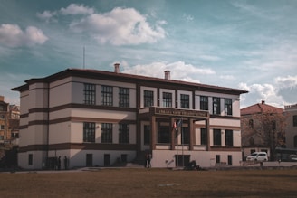 A large, historic building with a two-story structure adorned with symmetrical windows. The facade features white walls with brown accents and a sign displaying 'Ankara Vakıf Eserleri Müzesi' above the entrance. Two flags are visible at the main entrance, and several people are gathered near the building. The sky is partly cloudy, adding a dramatic backdrop to the scene.