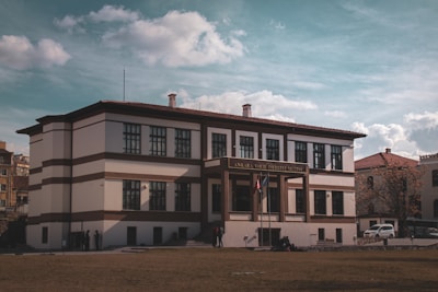 A large, historic building with a two-story structure adorned with symmetrical windows. The facade features white walls with brown accents and a sign displaying 'Ankara Vakıf Eserleri Müzesi' above the entrance. Two flags are visible at the main entrance, and several people are gathered near the building. The sky is partly cloudy, adding a dramatic backdrop to the scene.