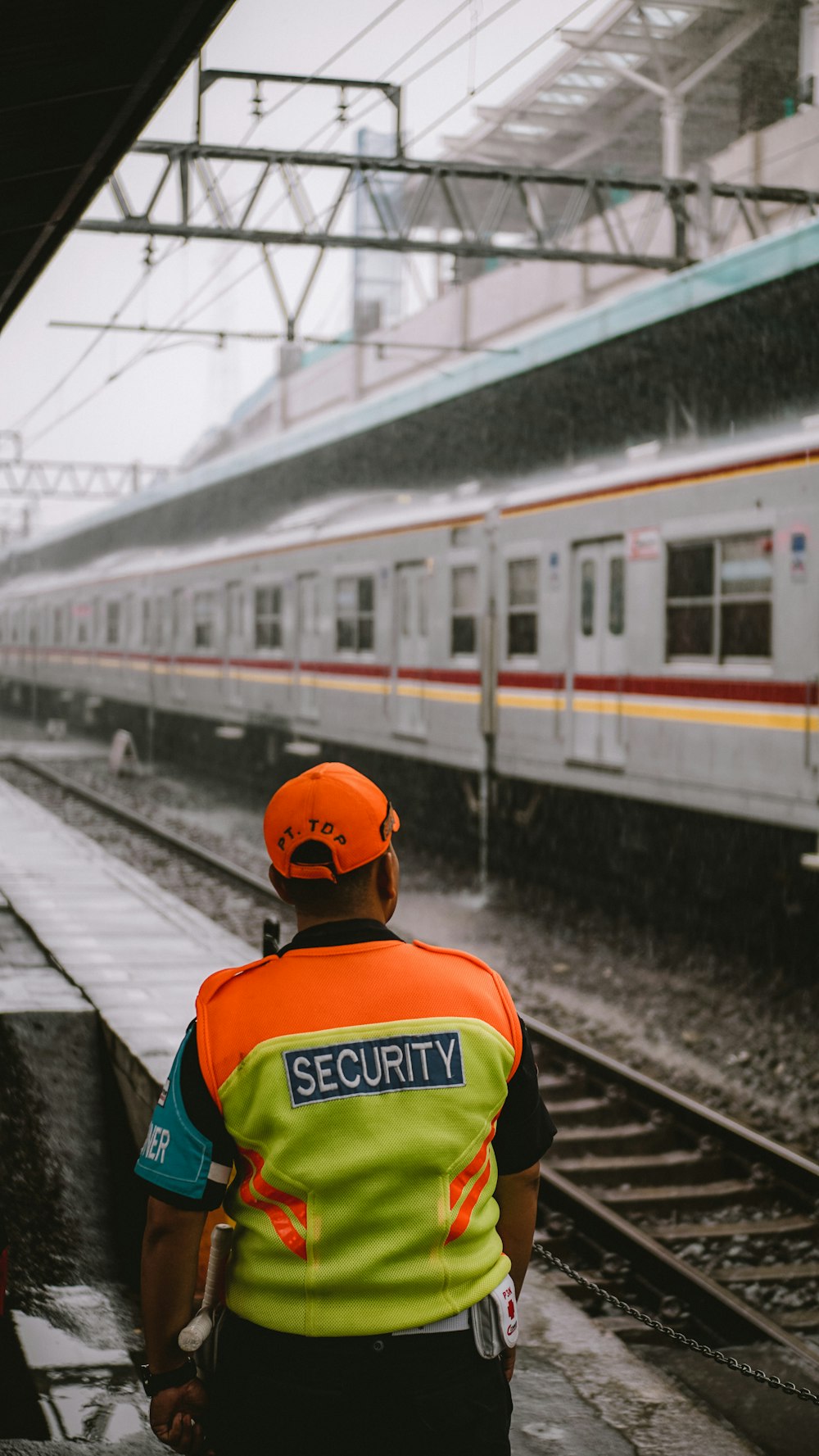 Hombre con camisa verde y amarilla con gorra naranja de pie en la estación de tren