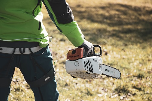 A person dressed in green workwear holds a chainsaw in their right hand. The background is a grassy area with some sunlight. The focus is on the lower body and the tool, suggesting activity related to landscaping or tree cutting.