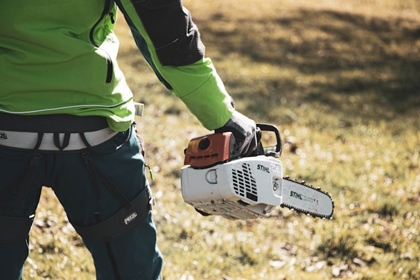 A person dressed in green workwear holds a chainsaw in their right hand. The background is a grassy area with some sunlight. The focus is on the lower body and the tool, suggesting activity related to landscaping or tree cutting.