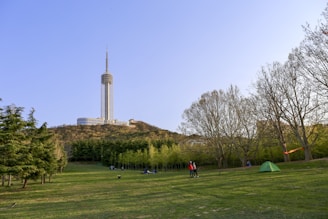 A tall communications tower stands on a hilltop surrounded by trees and a landscaped park area. A group of people is gathered on the grassy field, and a green tent is pitched nearby. The sky is clear and blue, suggesting a sunny day.