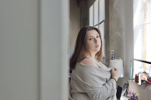 A plus size model wearing a beige knit cardigan, standing in a softly lit elegant room with natural light highlighting the texture of the fabric.