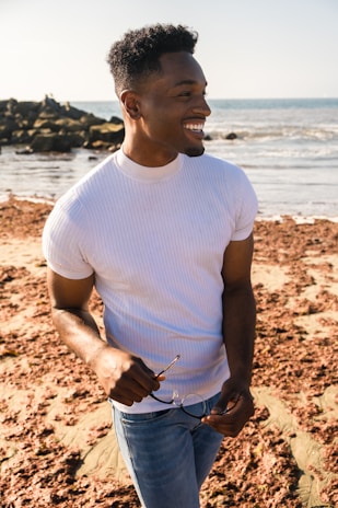 A smiling person wearing sunglasses and a sunhat, holding a bottle of Beach Block lotion under bright sunlight.