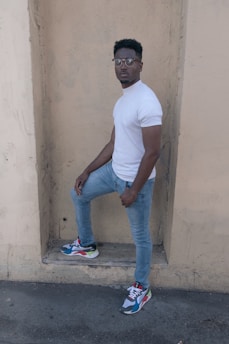 A person stands confidently against a beige, textured wall, wearing a white, short-sleeve shirt, light blue jeans, and colorful sneakers. The overall posture and expression suggest a casual, yet self-assured demeanor.