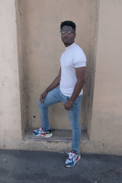A person stands confidently against a beige, textured wall, wearing a white, short-sleeve shirt, light blue jeans, and colorful sneakers. The overall posture and expression suggest a casual, yet self-assured demeanor.
