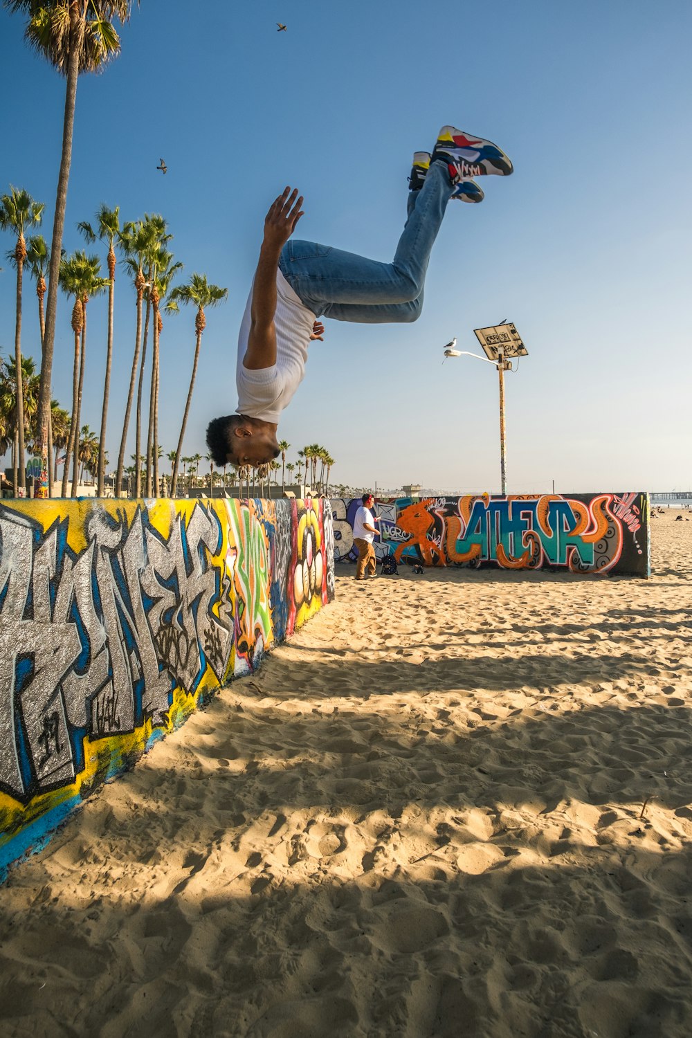 Uomo in maglietta bianca e pantaloni neri che fa acrobazie con lo skateboard sulla spiaggia durante il giorno