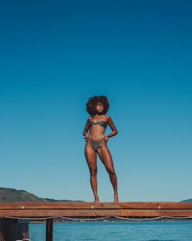 A woman stands confidently on a wooden dock above a clear body of water, wearing a two-piece swimsuit. The backdrop features a striking blue sky and distant hills, suggesting a serene, summer setting.