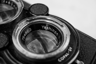 Close-up photo of the lens of an old camera with 'Yashinon' and a serial number engraved. The image is detailed, highlighting the metal and glass components, showcasing wear on the rim, indicating age.