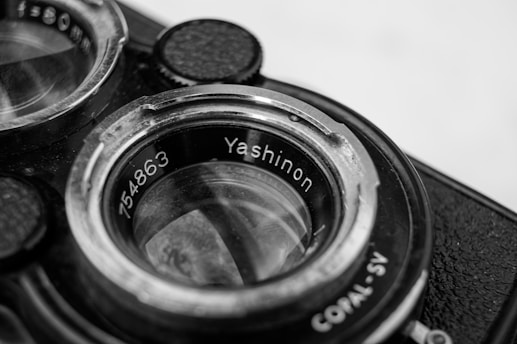 Close-up photo of the lens of an old camera with 'Yashinon' and a serial number engraved. The image is detailed, highlighting the metal and glass components, showcasing wear on the rim, indicating age.