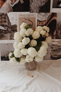 A bouquet of fluffy white flowers is arranged in a geometric vase, set against a background of black-and-white photographs and artwork. The foreground features a smooth white fabric surface with a gold hair clip and a pearl hair accessory placed on it.