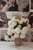 A geometric vase filled with large, white hydrangea blooms sits on a surface. Behind the vase is a wall adorned with various art prints and black-and-white photographs, including elements like a city scene, a beach, and the word 'Matisse'. The arrangement gives a vintage and artistic feel.