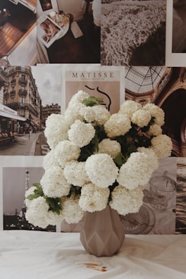 A geometric vase filled with large, white hydrangea blooms sits on a surface. Behind the vase is a wall adorned with various art prints and black-and-white photographs, including elements like a city scene, a beach, and the word 'Matisse'. The arrangement gives a vintage and artistic feel.