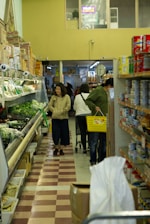 A grocery store aisle with several people shopping. On the left, there are shelves stocked with fresh vegetables and leafy greens. On the right, rows of canned goods and other packaged food items. The aisle has a tiled floor with a checkered pattern. A woman is pushing a shopping cart, and another person is carrying a yellow basket filled with groceries.