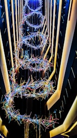 A vertical installation featuring intertwined loops of colorful lights suspended in a narrow, tall atrium with surrounding modern architecture and linear lighting.