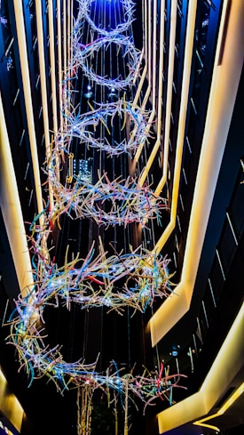 A vertical installation featuring intertwined loops of colorful lights suspended in a narrow, tall atrium with surrounding modern architecture and linear lighting.