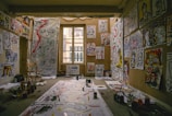 A room filled with abstract paintings covering the walls and floor. The paintings feature bold, expressive faces and colorful designs. Paints, brushes, and art supplies are scattered around. A wooden chair is positioned beside a large paper, suggesting an artist's workspace.