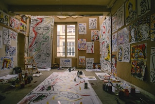 A room filled with abstract paintings covering the walls and floor. The paintings feature bold, expressive faces and colorful designs. Paints, brushes, and art supplies are scattered around. A wooden chair is positioned beside a large paper, suggesting an artist's workspace.