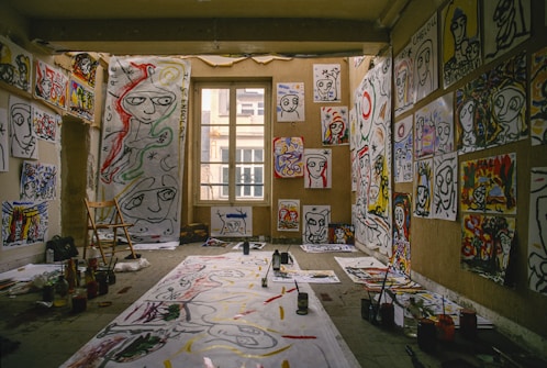 A room filled with abstract paintings covering the walls and floor. The paintings feature bold, expressive faces and colorful designs. Paints, brushes, and art supplies are scattered around. A wooden chair is positioned beside a large paper, suggesting an artist's workspace.