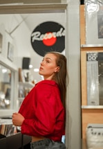 A woman is standing inside a record store, leaning against a wall. She is wearing a red jacket and denim pants. Her gaze is directed upwards, suggesting a contemplative or thoughtful expression. Vinyl records are visible in the background, along with a sign that partially reads 'Records' with a red circular design.