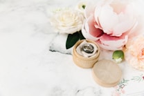 A wooden ring box containing a pair of rings is surrounded by soft pink and white flowers on a light marble surface. The scene includes roses, peonies, and a small green bud, conveying a romantic and elegant theme.