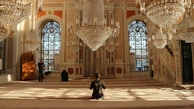 An opulent interior with ornate chandeliers cascading from the ceiling, grand arched windows, and intricate patterns on the walls. Sunlight filters through the windows, casting geometric shadows on the plush carpet. Individuals are seated or standing in contemplation or prayer, enhancing the serene and sacred atmosphere of the space.