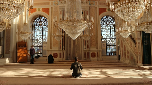 An opulent interior with ornate chandeliers cascading from the ceiling, grand arched windows, and intricate patterns on the walls. Sunlight filters through the windows, casting geometric shadows on the plush carpet. Individuals are seated or standing in contemplation or prayer, enhancing the serene and sacred atmosphere of the space.