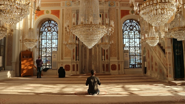 An opulent interior with ornate chandeliers cascading from the ceiling, grand arched windows, and intricate patterns on the walls. Sunlight filters through the windows, casting geometric shadows on the plush carpet. Individuals are seated or standing in contemplation or prayer, enhancing the serene and sacred atmosphere of the space.