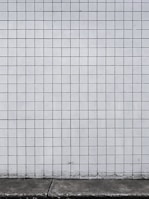 A grid of white tiles covers a wall with a concrete base at the bottom. The tiles are arranged in a uniform pattern with dark grout lines that create a distinct grid structure.