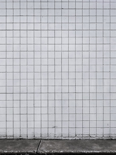 A grid of white tiles covers a wall with a concrete base at the bottom. The tiles are arranged in a uniform pattern with dark grout lines that create a distinct grid structure.