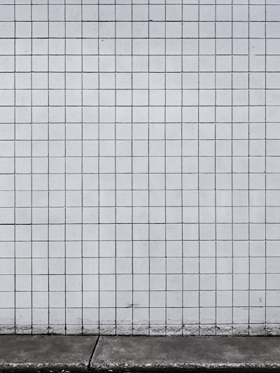 A grid of white tiles covers a wall with a concrete base at the bottom. The tiles are arranged in a uniform pattern with dark grout lines that create a distinct grid structure.