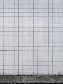 A grid of white tiles covers a wall with a concrete base at the bottom. The tiles are arranged in a uniform pattern with dark grout lines that create a distinct grid structure.