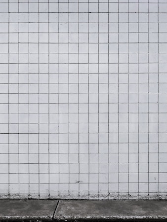 A grid of white tiles covers a wall with a concrete base at the bottom. The tiles are arranged in a uniform pattern with dark grout lines that create a distinct grid structure.