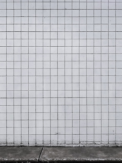 A grid of white tiles covers a wall with a concrete base at the bottom. The tiles are arranged in a uniform pattern with dark grout lines that create a distinct grid structure.