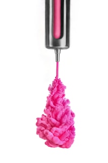 Vibrant pink ink is being injected from a metallic syringe-like device into a clear liquid, creating swirling patterns and clouds.