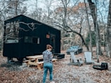 A person stands near a black tiny house in the woods, surrounded by autumn trees. Nearby, there is a wooden picnic table and several Adirondack chairs positioned around a fire pit. A car is parked close by on the gravel path.