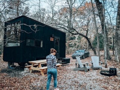 A person stands near a black tiny house in the woods, surrounded by autumn trees. Nearby, there is a wooden picnic table and several Adirondack chairs positioned around a fire pit. A car is parked close by on the gravel path.