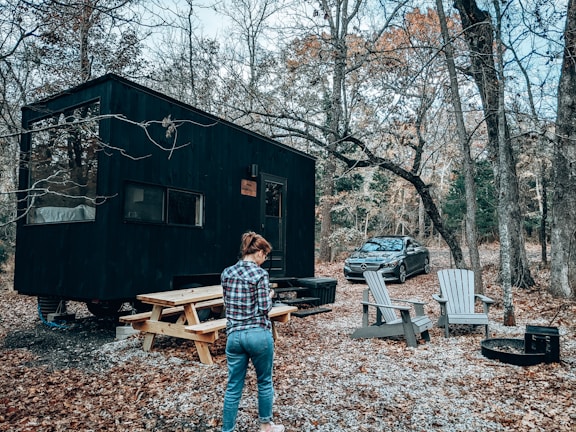 A person stands near a black tiny house in the woods, surrounded by autumn trees. Nearby, there is a wooden picnic table and several Adirondack chairs positioned around a fire pit. A car is parked close by on the gravel path.