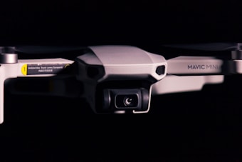 A close-up view of a drone against a dark background. The drone has a camera on the front and visible text that reads 'Mavic Mini'. One of the arms has a yellow and black label with instructions for unfolding.