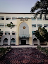A tall, multi-story building with a prominent entrance decorated in an architectural style featuring geometric patterns. The sign 'Dentistry' is displayed above the entrance. The building is surrounded by a few trees and neatly trimmed bushes. A tiled pathway leads up to the entrance, and a traffic cone is placed near the doorway.