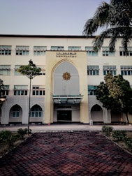 A tall, multi-story building with a prominent entrance decorated in an architectural style featuring geometric patterns. The sign 'Dentistry' is displayed above the entrance. The building is surrounded by a few trees and neatly trimmed bushes. A tiled pathway leads up to the entrance, and a traffic cone is placed near the doorway.