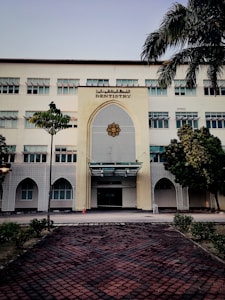 A tall, multi-story building with a prominent entrance decorated in an architectural style featuring geometric patterns. The sign 'Dentistry' is displayed above the entrance. The building is surrounded by a few trees and neatly trimmed bushes. A tiled pathway leads up to the entrance, and a traffic cone is placed near the doorway.