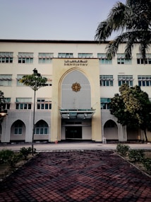 A tall, multi-story building with a prominent entrance decorated in an architectural style featuring geometric patterns. The sign 'Dentistry' is displayed above the entrance. The building is surrounded by a few trees and neatly trimmed bushes. A tiled pathway leads up to the entrance, and a traffic cone is placed near the doorway.