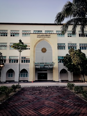 A tall, multi-story building with a prominent entrance decorated in an architectural style featuring geometric patterns. The sign 'Dentistry' is displayed above the entrance. The building is surrounded by a few trees and neatly trimmed bushes. A tiled pathway leads up to the entrance, and a traffic cone is placed near the doorway.