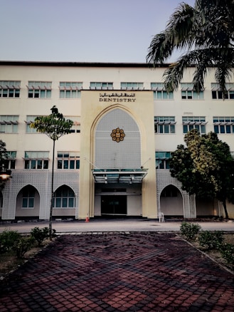 A tall, multi-story building with a prominent entrance decorated in an architectural style featuring geometric patterns. The sign 'Dentistry' is displayed above the entrance. The building is surrounded by a few trees and neatly trimmed bushes. A tiled pathway leads up to the entrance, and a traffic cone is placed near the doorway.
